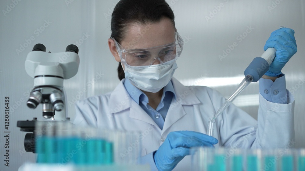 Scientist woman wearing a lab coat, mask, and safety glasses is using a micropipette to drop a blue liquid into a test tube, portrait view. Medicine and science concept Scientist woman wearing a lab coat, mask, and safety glasses is using a micropipette to drop a blue liquid into a test tube, portrait view. Medicine and science concept