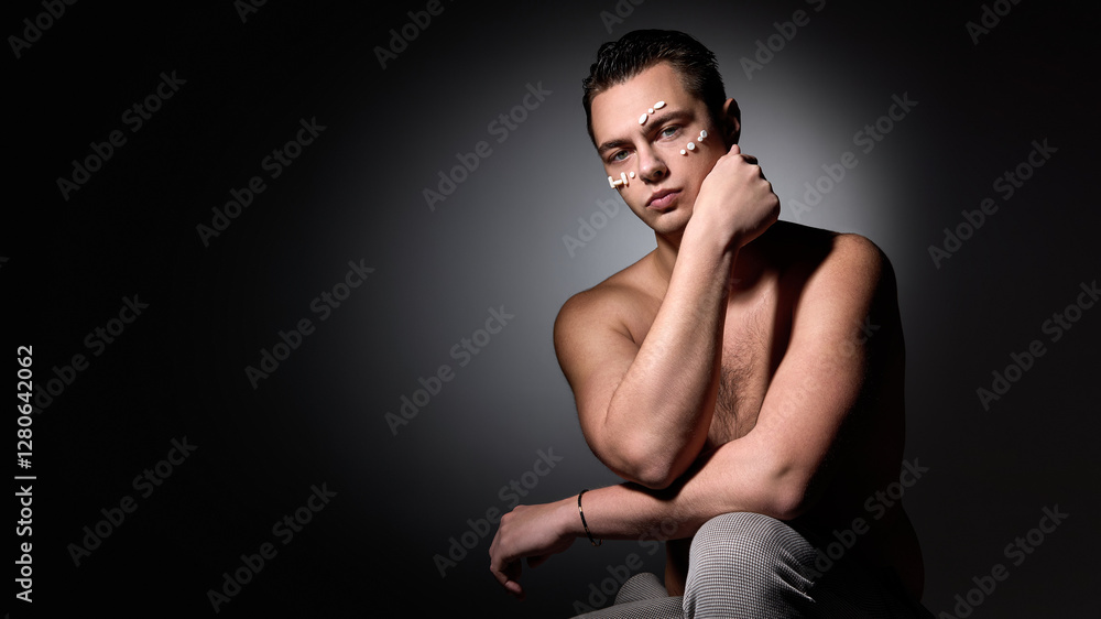 Naklejka premium Shirtless young man sitting with arm resting on knee, white pills on face, looking into camera with moody lighting against dark studio background. Concept of selfcare, natural beauty, cosmetology.
