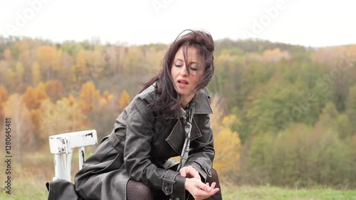 Autumn landscape with gothic woman on chair. Stylish woman and chair in autumn landscape outdoor. Fall nature landscape and girl. Central park in New York. Girl in fall trench. Sitting on a hillto