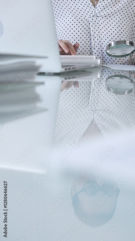 Businesswoman in white dotted blouse is carefully reviewing financial reports with magnifying glass and calculator, conducting audit and analyzing data, vertical view Taxes, audit in business