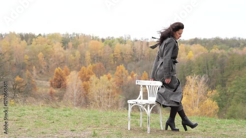 Autumn landscape with gothic woman on chair, copy space. Stylish woman and chair in autumn landscape outdoor. Fall nature landscape girl. Central park in New York. Girl in fall trench. Slow motion.