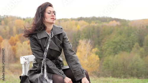 Autumn landscape with gothic woman on chair, copy space. Stylish woman chair in autumn landscape outdoor. Fall nature landscape and girl. Central park in New York. Girl in fall trench. Strong wind.