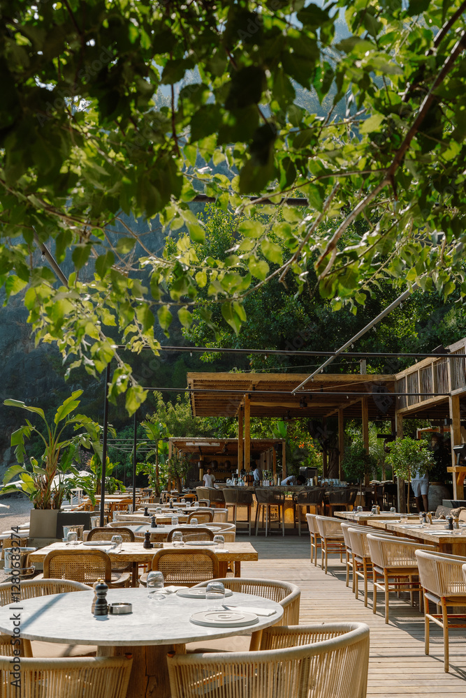 Fototapeta premium A stylish open-air restaurant in Fethiye, Türkiye, with wooden tables, wicker chairs, and lush greenery, creating a relaxed dining atmosphere under natural shade in a scenic coastal setting
