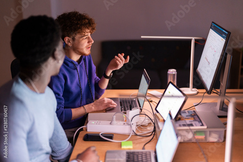 Two young men sit at a desk in a dimly lit office, discussing financial data displayed on multiple screens while analyzing stock market trends and investment opportunities.