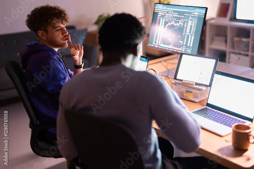 A young man in a blue hoodie eats a sandwich while another man works at a desk with multiple screens displaying stock market data and digital charts.