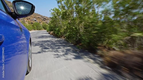 A stunning side-mounted camera shot from a moving car, capturing the vibrant blue vehicle, winding roads, lush green bushes, and the rugged Mediterranean terrain of Crete, Greece.