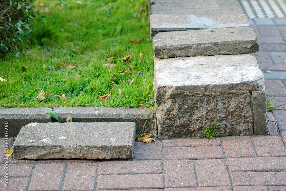 Damaged curb with displaced concrete blocks on brick walkway, need for repair and maintenance in public spaces. Concrete curb separating grassy area from sidewalk has broken blocks, neglect and wear.