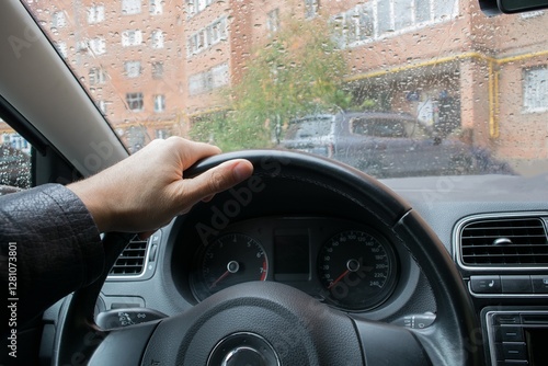 One hand of the driver is on the steering wheel of the car when driving in the yard during the rain.