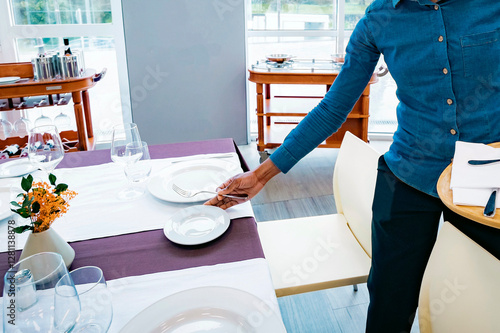 Waiter arranging an elegant table in a bright restaurant. Fine dining setup with glassware, plates, and decoration for a high-end dining experience. photo