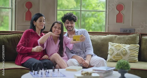 Indian Asian happy mom lady with young adult child sitting cozy sofa couch use mobile take selfie indoor home cute teen girl boy hold phone look cam make funny pose have fun joy enjoy day spend time