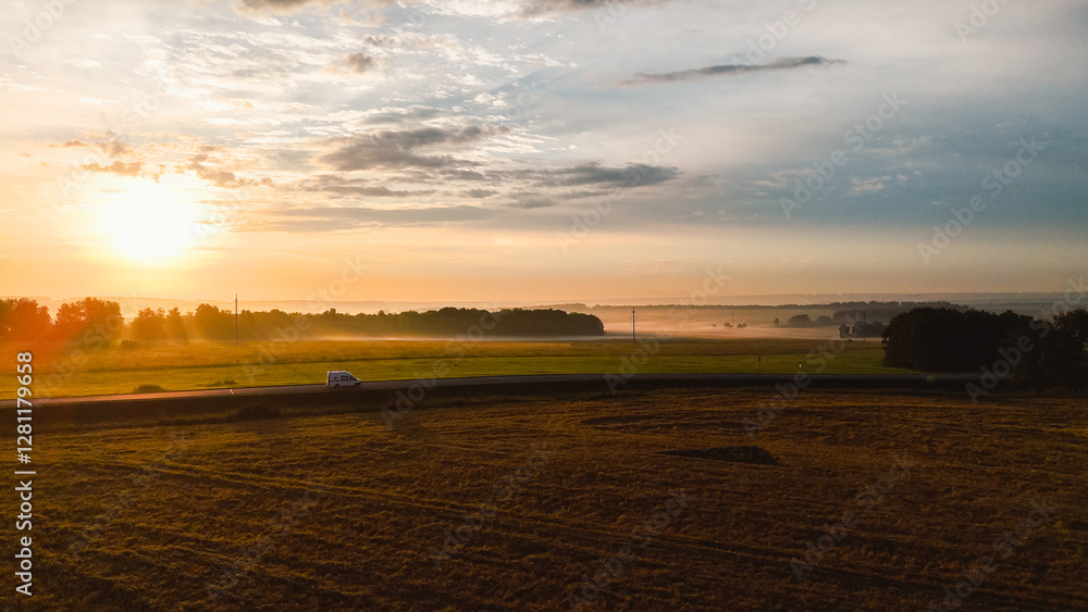 Fototapeta premium dawn in a wheat field, a car is driving along the highway, fog is creeping on the horizon