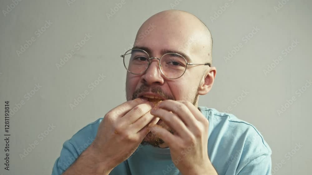A bald man sloppily pushes a large piece of burger into his mouth, trying to gorge himself. He happily licks his fingers, stained with barbecue sauce, savoring the unhealthful food. A bald man sloppily pushes a large piece of burger into his mouth, trying to gorge himself. He happily licks his fingers, stained with barbecue sauce, savoring the unhealthful food.