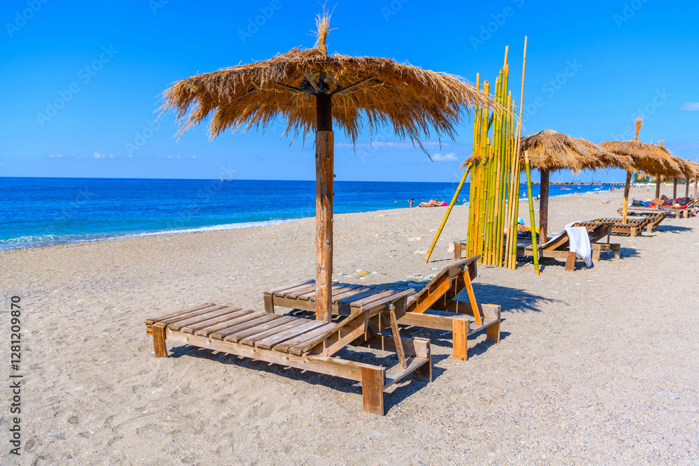 Straw beach umbrella with sun loungers in a row on the sandy beach of the sea in Gagra, Abkhazia.Trips, vacation travel, relaxation on the ocean coast in a tropical country. Copy space. Straw beach umbrella with sun loungers in a row on the sandy beach of the sea in Gagra, Abkhazia.Trips, vacation travel, relaxation on the ocean coast in a tropical country. Copy space.