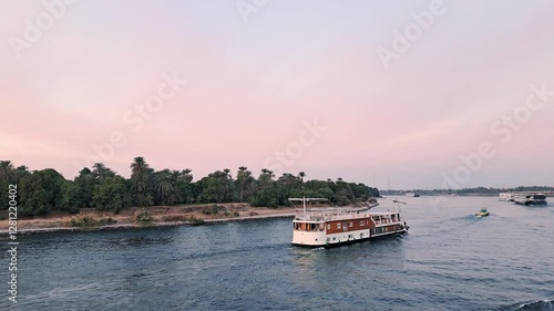 Evening scene on the Nile, featuring a cruise boat navigating the river as the sun sets. Multiple boats dot the waterway under a soft, pink-tinged sky. Egypt.