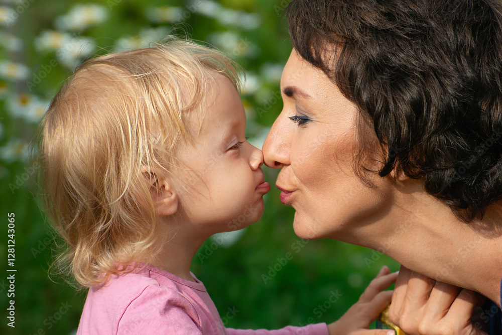 Mother and child rub noses. A funny little girl kisses her mother on the lips. Close-up of cheerful mother and baby nose to nose in profile against chamomile background. Happy motherhood concept. Mother and child rub noses. A funny little girl kisses her mother on the lips. Close-up of cheerful mother and baby nose to nose in profile against chamomile background. Happy motherhood concept.
