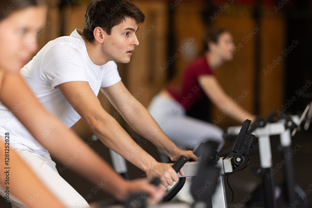 Guy hardly performs exercises on stationary bicycle during fitness class. Gym and fitness equipment, visitors are engaged in physical exercises during training Guy hardly performs exercises on stationary bicycle during fitness class. Gym and fitness equipment, visitors are engaged in physical exercises during training