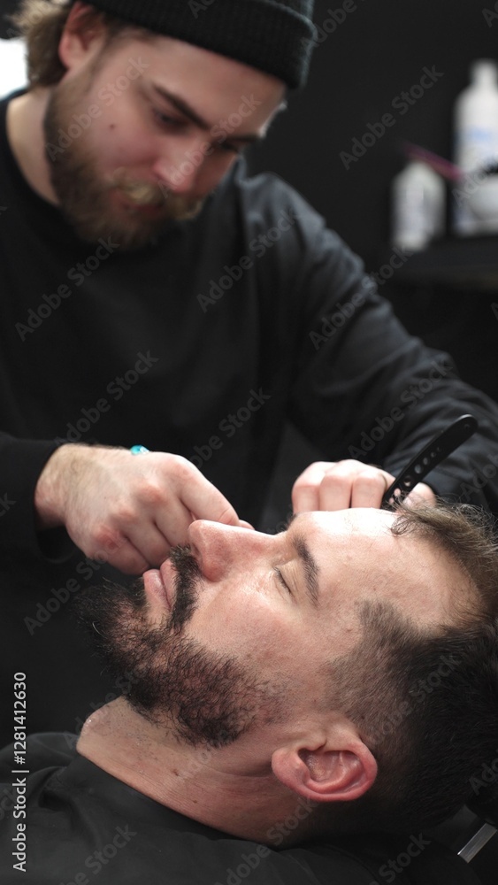 Fototapeta premium Professional barber carefully shaving client's beard with straight razor, applying gel and using hot towel for precise grooming in contemporary barbershop setting, vertical view