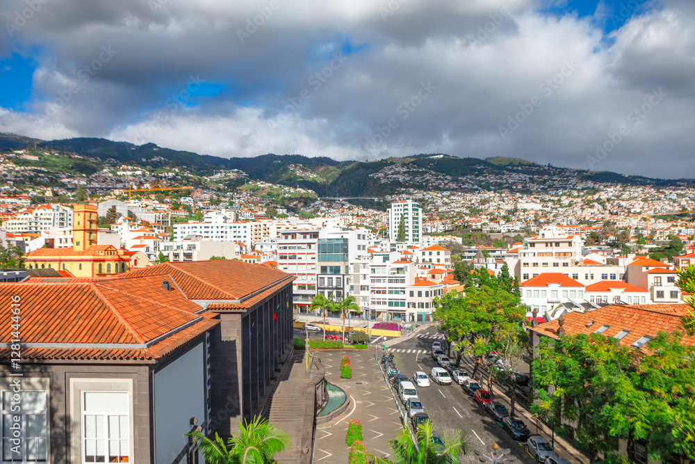Panoramic view of Funchal, Madeira, with terracotta roofed buildings, lush green hills and vibrant cityscape under partly cloudy sky. Mix of modern and traditional architecture highlights island charm