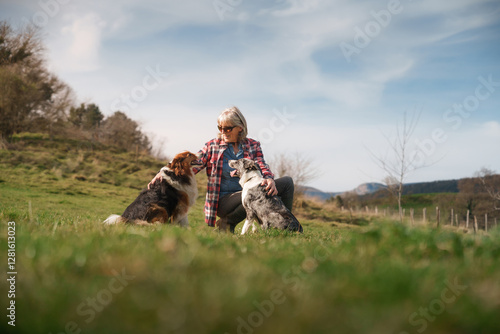 Female farmer crouching in grass field petting two dogs. Senior woman farmer petting her two border collies in a green field