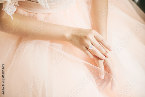 Close-up of a bride’s hands resting gently on a soft blush tulle gown, showcasing a sparkling diamond engagement ring. The delicate lace details and pastel tones add a romantic and elegant touch. photo