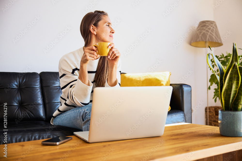 Woman enjoying cup of coffee while working on laptop at home sitting on sofa Woman enjoying cup of coffee while working on laptop at home sitting on sofa