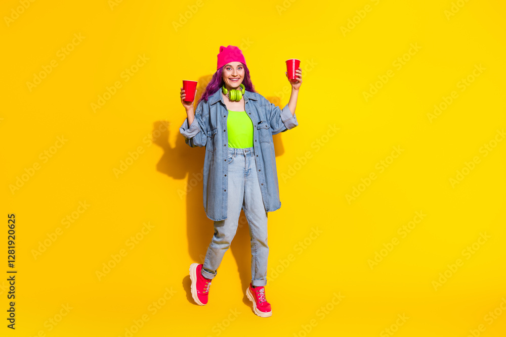 Stylish woman in casual denim outfit on vibrant yellow background holding red cups and expressing a joyful vibe Stylish woman in casual denim outfit on vibrant yellow background holding red cups and expressing a joyful vibe