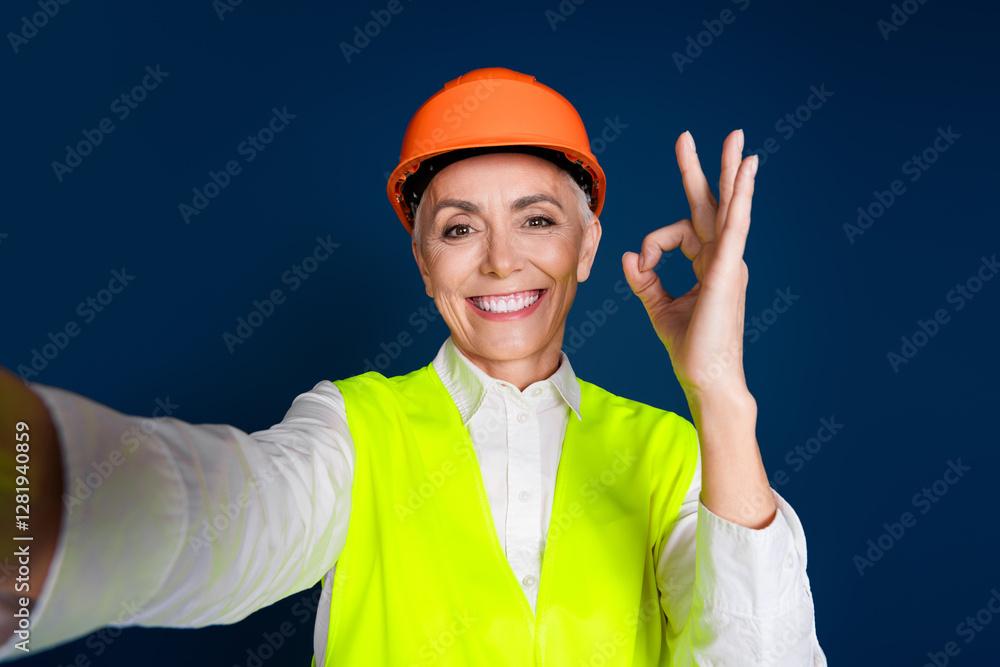 Confident senior woman engineer making OK gesture while wearing a high-visibility vest and hardhat against a dark blue background Confident senior woman engineer making OK gesture while wearing a high-visibility vest and hardhat against a dark blue background