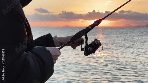 Slim girl wearing black spins the reel of a fishing rod on the calm coast, with sun setting behind clouds in the background. Close-up