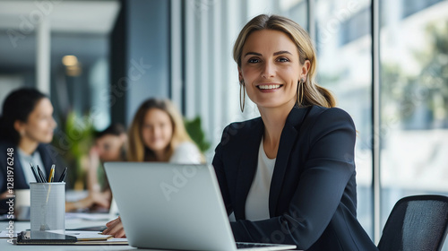 Confident businesswoman in professional attire, smiling at her desk with a laptop while team members collaborate in a modern office with bright lighting, large windows, and a productive atmosphere.