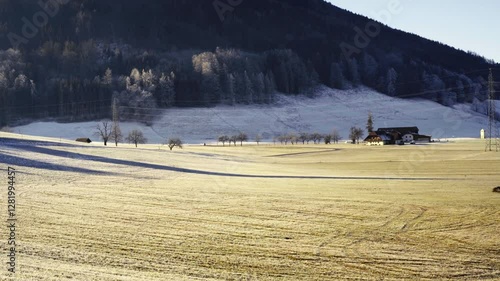 Scenic winter landscape with frost-covered fields, a farmhouse, and distant forested hills under clear blue skies on a crisp morning.
