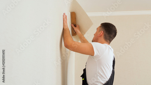 Vinyl wallpapers squeegee tool in use by professional interior renovation worker. Young man with dark hair in a uniform glueing beige wallpaper at home. photo