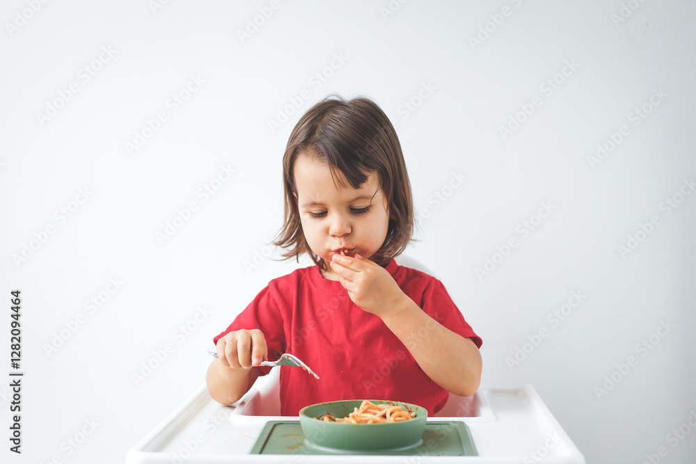 Little girl with brown hair sitting in a high chair against a white background, wearing a red shirt, eating spaghetti with both hands and a spoon. Her face is slightly messy as she enjoys her meal Little girl with brown hair sitting in a high chair against a white background, wearing a red shirt, eating spaghetti with both hands and a spoon. Her face is slightly messy as she enjoys her meal