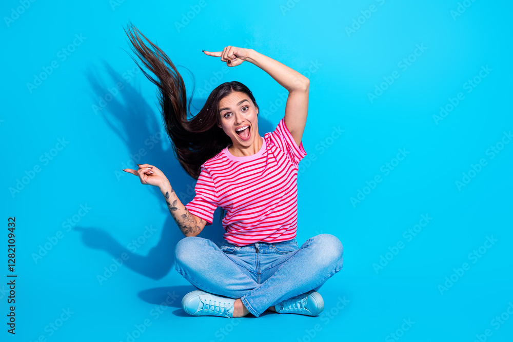 Happy young woman gesturing energetically while sitting on a bright blue background wearing a striped pink t-shirt and casual jeans Happy young woman gesturing energetically while sitting on a bright blue background wearing a striped pink t-shirt and casual jeans