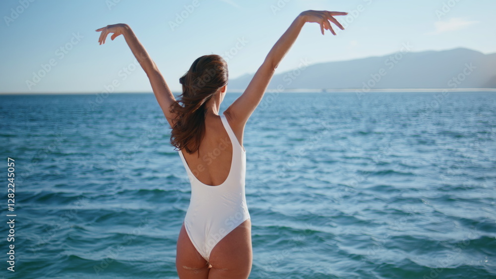 Swimsuit woman raising hands looking on ocean waves back view. Relaxed brunette Swimsuit woman raising hands looking on ocean waves back view. Relaxed brunette