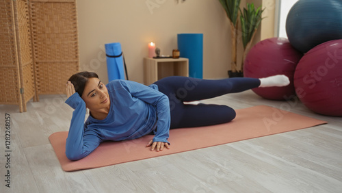 Young woman doing exercise on mat indoors at gym center, dressed in blue, surrounded by gym equipment like exercise balls and a candle, appearing focused and relaxed. photo