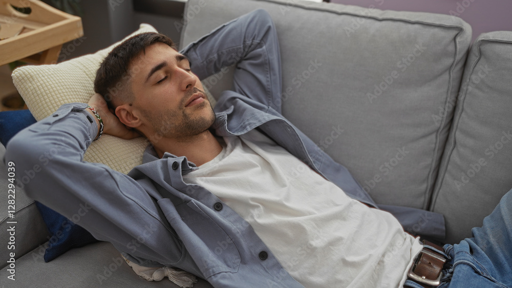 Fototapeta premium Young man relaxing on a sofa indoors with his arms behind his head, sporting casual attire, conveying a sense of comfort and tranquility in a modern home setting.