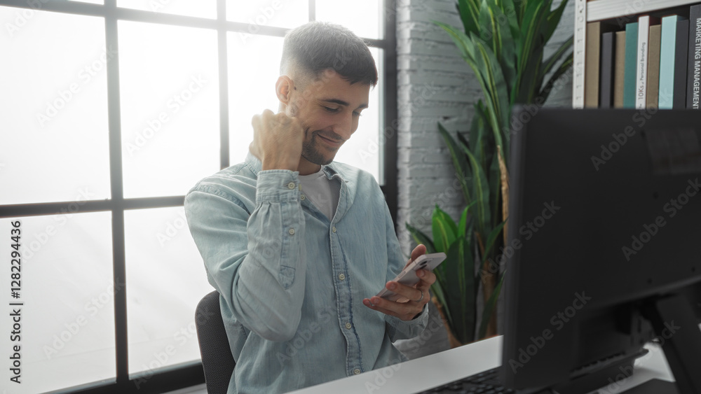 Young man checking smartphone in modern office with plants, expressing success with fist pump, captured from side showing computer monitor and books in background Young man checking smartphone in modern office with plants, expressing success with fist pump, captured from side showing computer monitor and books in background