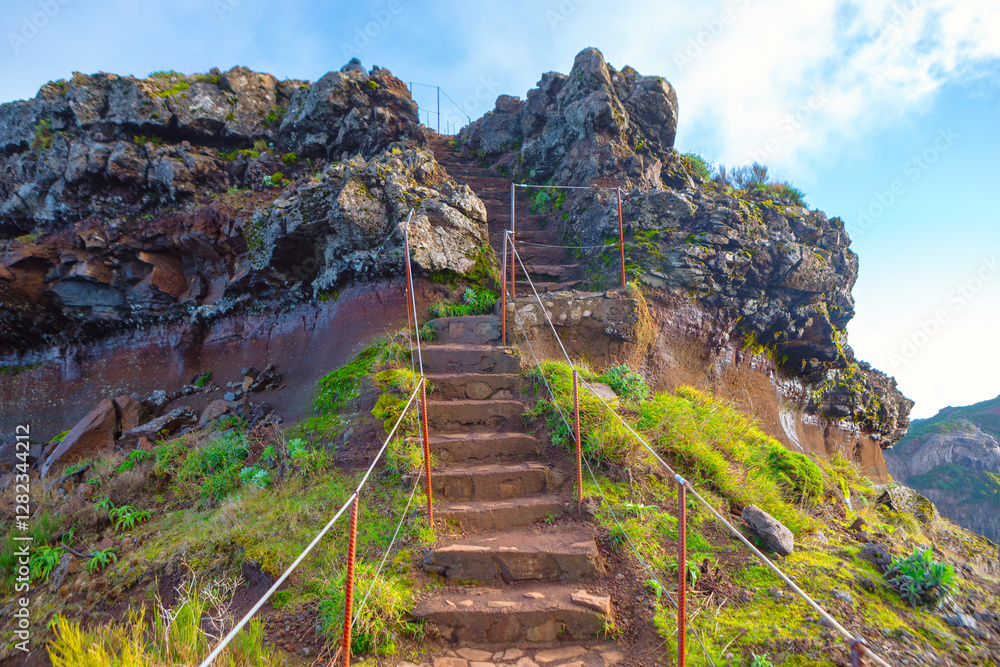 Stone steps leading up a rocky hill, with railing on both sides for support. Steps are surrounded by green vegetation, and the sky is partly cloudy, creating a scenic and inviting path for hikers