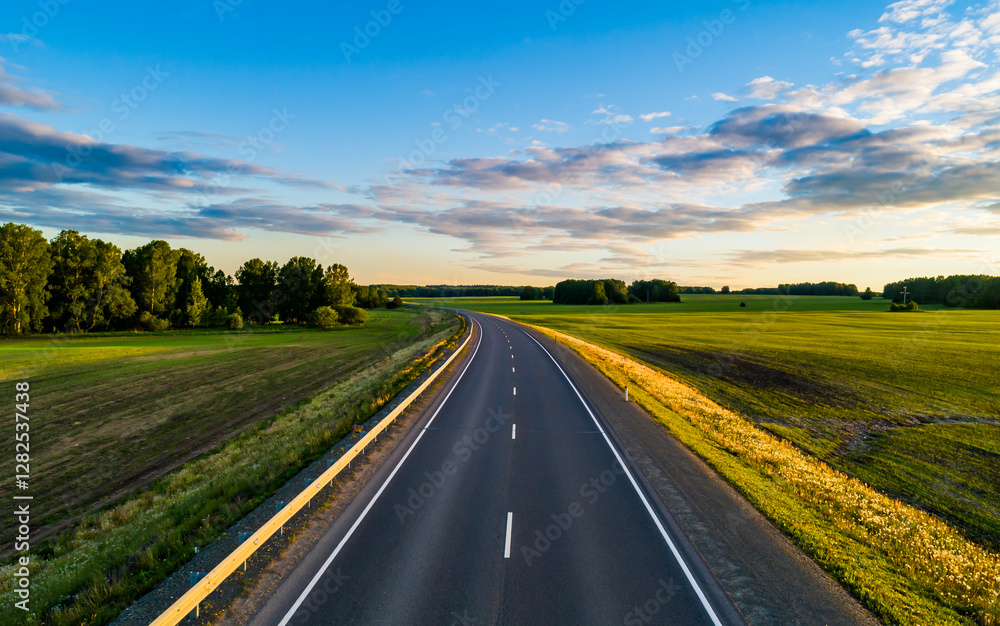 A winding road stretches into the distance, flanked by vibrant green fields and dense trees under a clear blue sky. A winding road stretches into the distance, flanked by vibrant green fields and dense trees under a clear blue sky.