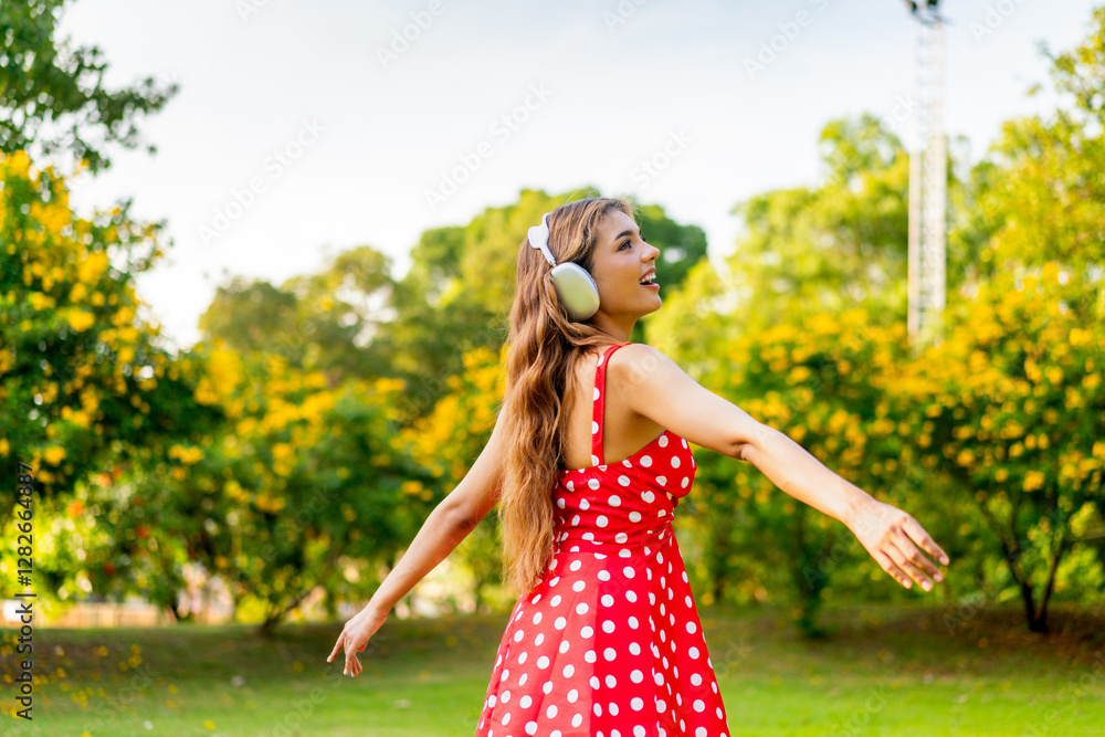 Obraz premium Young Woman in Retro Red Polka Dot Dress Wearing Headphones Listening and Enjoying Music in a Park Happily, Immersing with Nature
