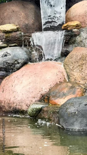 A serene waterfall cascading over smooth,natural looking rocks into a calm pond.The earthy tonesof the stones contrast beautifully with the flowing water,creating a peaceful anbd refreshing ambiance. photo