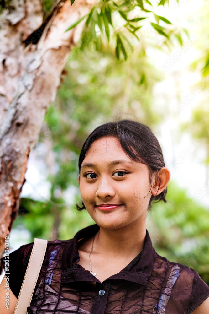 Portrait of a beautiful Asian teenage girl in casual stylish outfit standing near a tree, looking at the camera and smiling, with sky and greenery background Portrait of a beautiful Asian teenage girl in casual stylish outfit standing near a tree, looking at the camera and smiling, with sky and greenery background