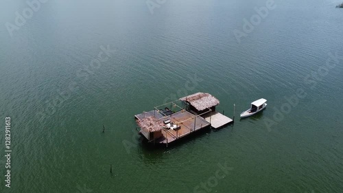 A floating bamboo hut with thatched roofs on Lake Tondano, Minahasa, Indonesia. A small boat is moored nearby, surrounded by calm, greenish water under a clear sky