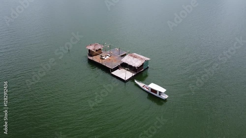 A floating bamboo hut with thatched roofs on Lake Tondano, Minahasa, Indonesia. A small boat is moored nearby, surrounded by calm, greenish water under a clear sky