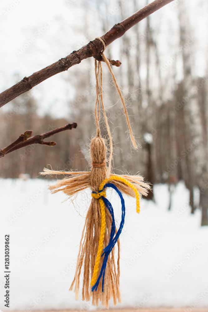 Knot doll with Ukrainian national colours hanging on a tree in winter park. Traditional handcraft amulet. Faceless straw raw with yellow and blue ribbons. Heritage art of Ukraine. Knot puppet.