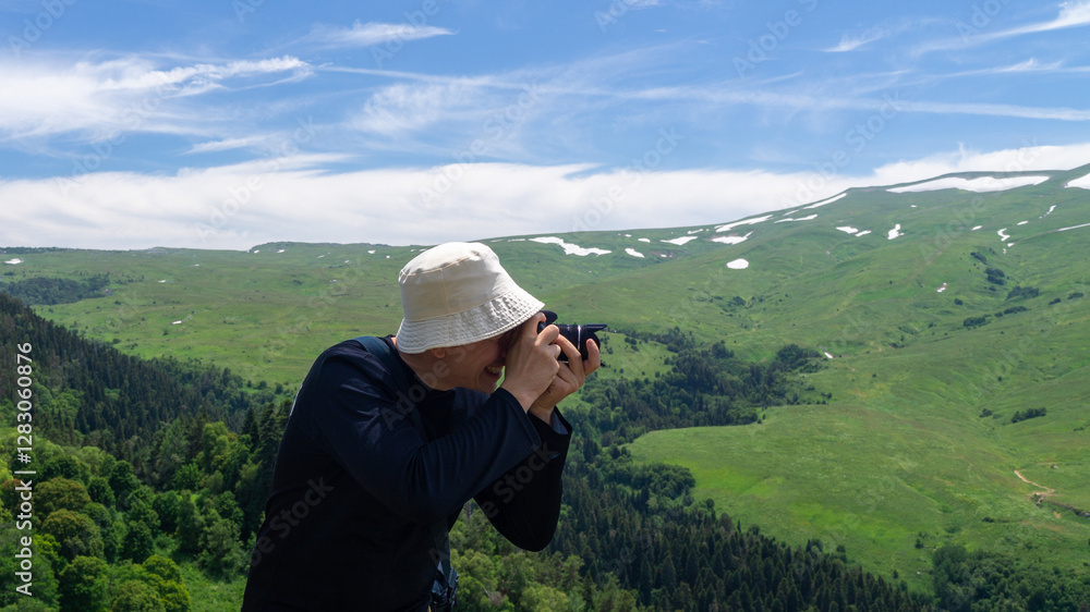 Young man taking landscape photo with DSLR camera on top of mountain in summer against green plateau Young man taking landscape photo with DSLR camera on top of mountain in summer against green plateau