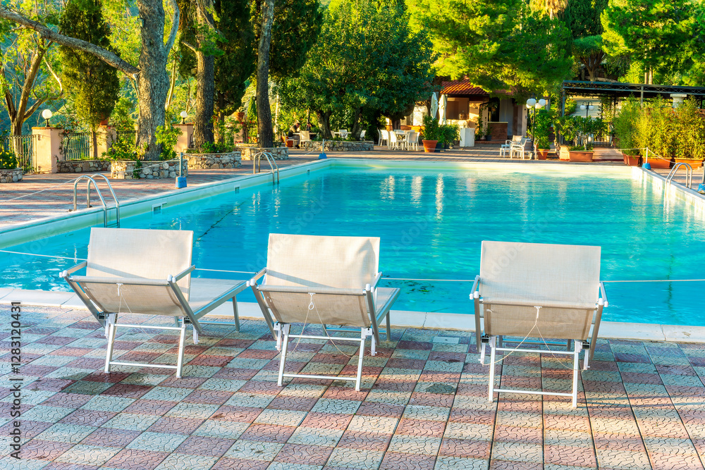 Fototapeta premium three white desk chairs or chaise lounge with blue swimming pool and hotel green garden on backgeound of vacation summer landscape