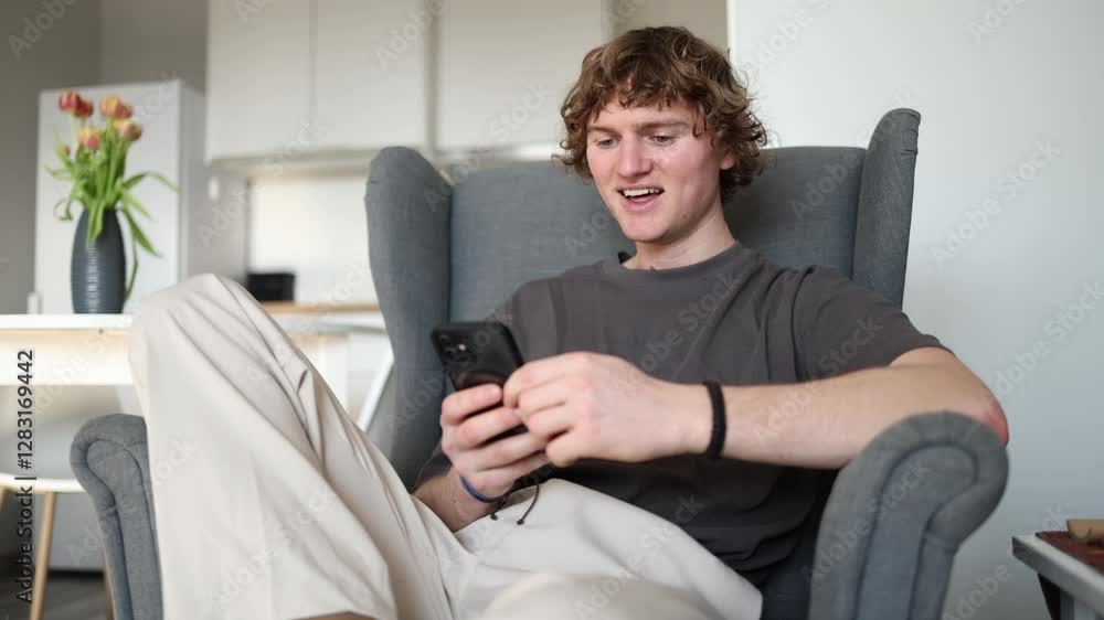 The young man seems happy and relaxed while looking at his phone, likely reading something amusing or engaging. His smile and relaxed posture show that he is enjoying the moment. The young man seems happy and relaxed while looking at his phone, likely reading something amusing or engaging. His smile and relaxed posture show that he is enjoying the moment.
