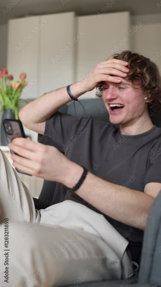 Vertical screen: Young man laughing hard at a funny social media video on his smartphone, joyful and amused emotions, casual leisure time at home, cozy atmosphere in modern living room. Vertical screen: Young man laughing hard at a funny social media video on his smartphone, joyful and amused emotions, casual leisure time at home, cozy atmosphere in modern living room.