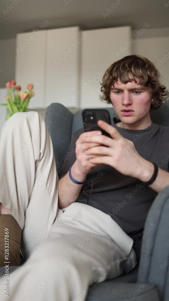 Vertical screen: Pleasantly surprised young man looking intrigued at his smartphone while sitting relaxed on sofa at home, positive emotional reaction, leisure activity, modern cozy interior. Vertical screen: Pleasantly surprised young man looking intrigued at his smartphone while sitting relaxed on sofa at home, positive emotional reaction, leisure activity, modern cozy interior.